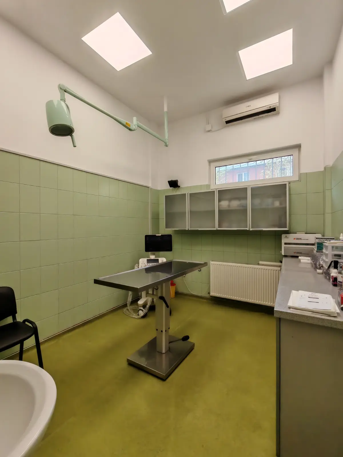 Sala de consultații: masă de examinare din inox, lampă articulată deasupra și ecograf lângă perete.